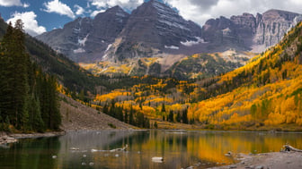 The twin Maroon Bells peaks, lightly dusted with snow, rise above hillsides of golden autumn aspen and dark evergreens, mirrored in the calm water of Maroon Lake under a blue sky with white clouds, Colorado.